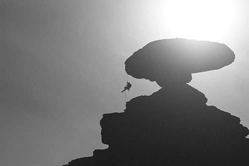 A black and white image of a climber rappelling from a mushroom shaped rock in the desert.